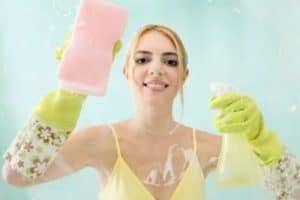 Pracująca kobieta sprzątająca okno z mopem i sprayem, uśmiechnięta, w ż&oacute;łtej bluzce i rękawicach.