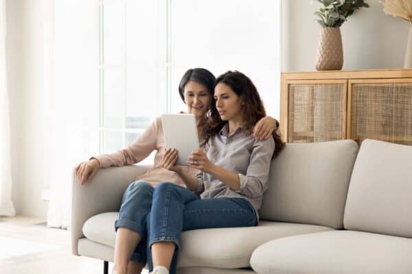 Jak przechowywać zdjęcia na urządzeniu - poradnik Dwie dorosłe kobiety – mama i córka – siedzą na kanapie i wspólnie przeglądają rodzinną galerię zdjęć przechowywaną w chmurze.
