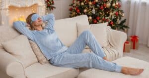Relaksująca kobieta na kanapie z opaską na oczach, obok bożonarodzeniowa dekoracja, idealne na domowe odprężenie.